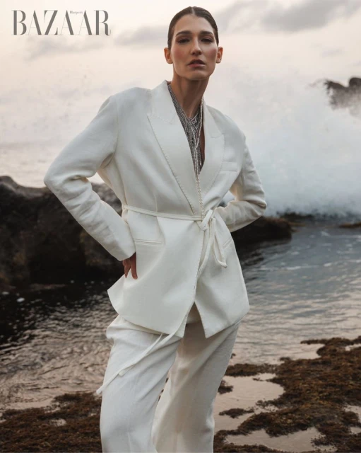 Editorial fashion shot of a woman in a white suit posing on a rocky beach with crashing waves in the background, neutral lighting.