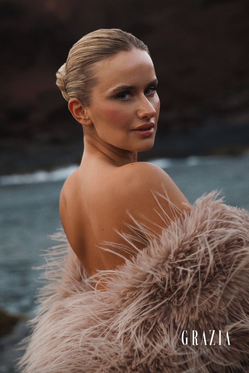 Blonde woman in feathered coat on beach for Grazia magazine, looking over her shoulder, with a blurred ocean background.