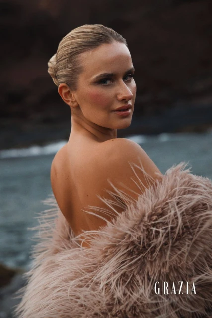 Blonde woman in feathered coat on beach for Grazia magazine, looking over her shoulder, with a blurred ocean background.