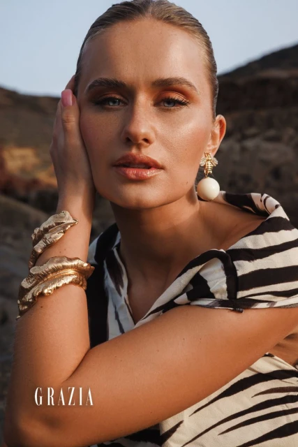 Close-up portrait of a blonde woman wearing gold bracelets, a zebra-striped dress, and bee earrings, posing outdoors against a rocky background.
