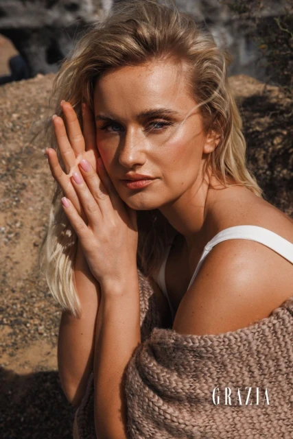 Close-up of a blonde woman in a desert setting, draped in a brown knit shawl, with her hands near her face, and the Grazia logo.