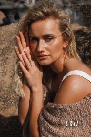 Close-up of a blonde woman in a desert setting, draped in a brown knit shawl, with her hands near her face, and the Grazia logo.
