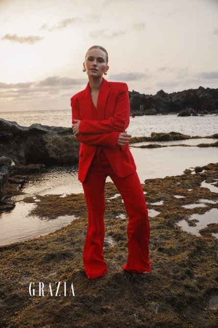 Woman in a red suit poses on a rocky beach with the ocean and cloudy sky in the background for a fashion editorial shoot.