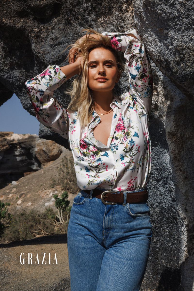 Blonde woman poses in a floral shirt and blue jeans against a rock formation; outdoor fashion editorial with natural light.