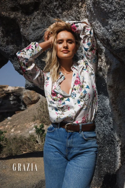 Blonde woman poses in a floral shirt and blue jeans against a rock formation; outdoor fashion editorial with natural light.