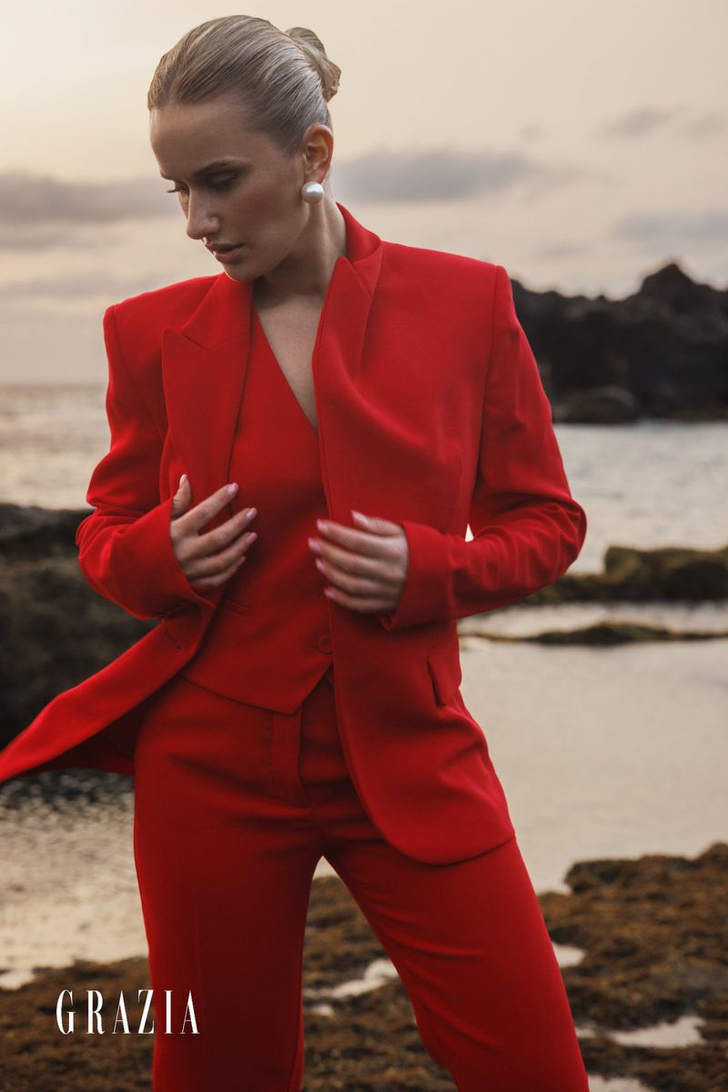 Fashion editorial portrait of a blonde female adult in a red three-piece suit, posing on a rocky beach with the ocean in the background.