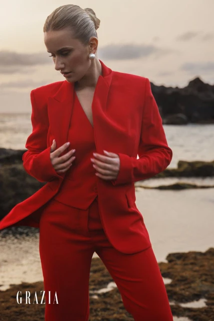 Fashion editorial portrait of a blonde female adult in a red three-piece suit, posing on a rocky beach with the ocean in the background.