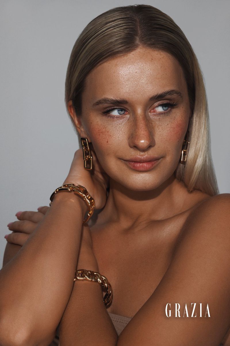 Portrait of a blond woman wearing gold chain earrings and matching bracelets, posing in a photo studio in soft, directional lighting.