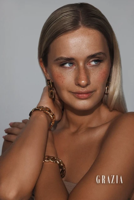 Portrait of a blond woman wearing gold chain earrings and matching bracelets, posing in a photo studio in soft, directional lighting.