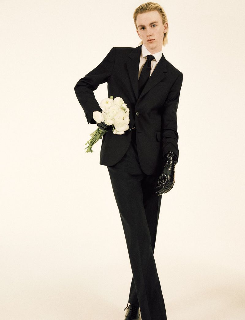 Man in a black suit and gloves holds white flowers on a beige background, fashion-forward formal wear.