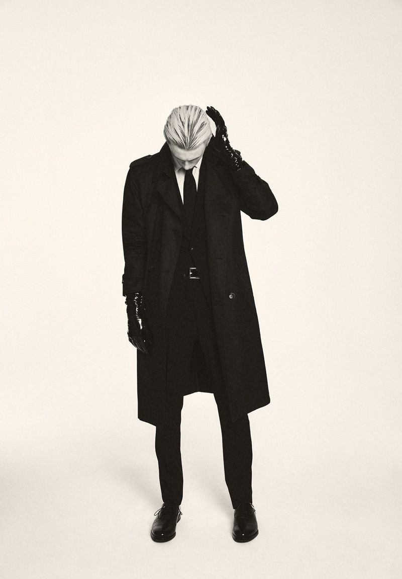 A slender adult male wearing a long black overcoat and gloves stands in a photo studio with a neutral expression and downward gaze.