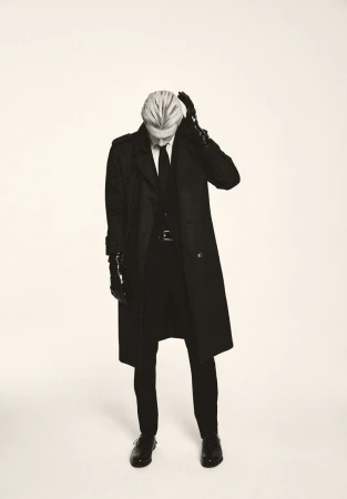 A slender adult male wearing a long black overcoat and gloves stands in a photo studio with a neutral expression and downward gaze.