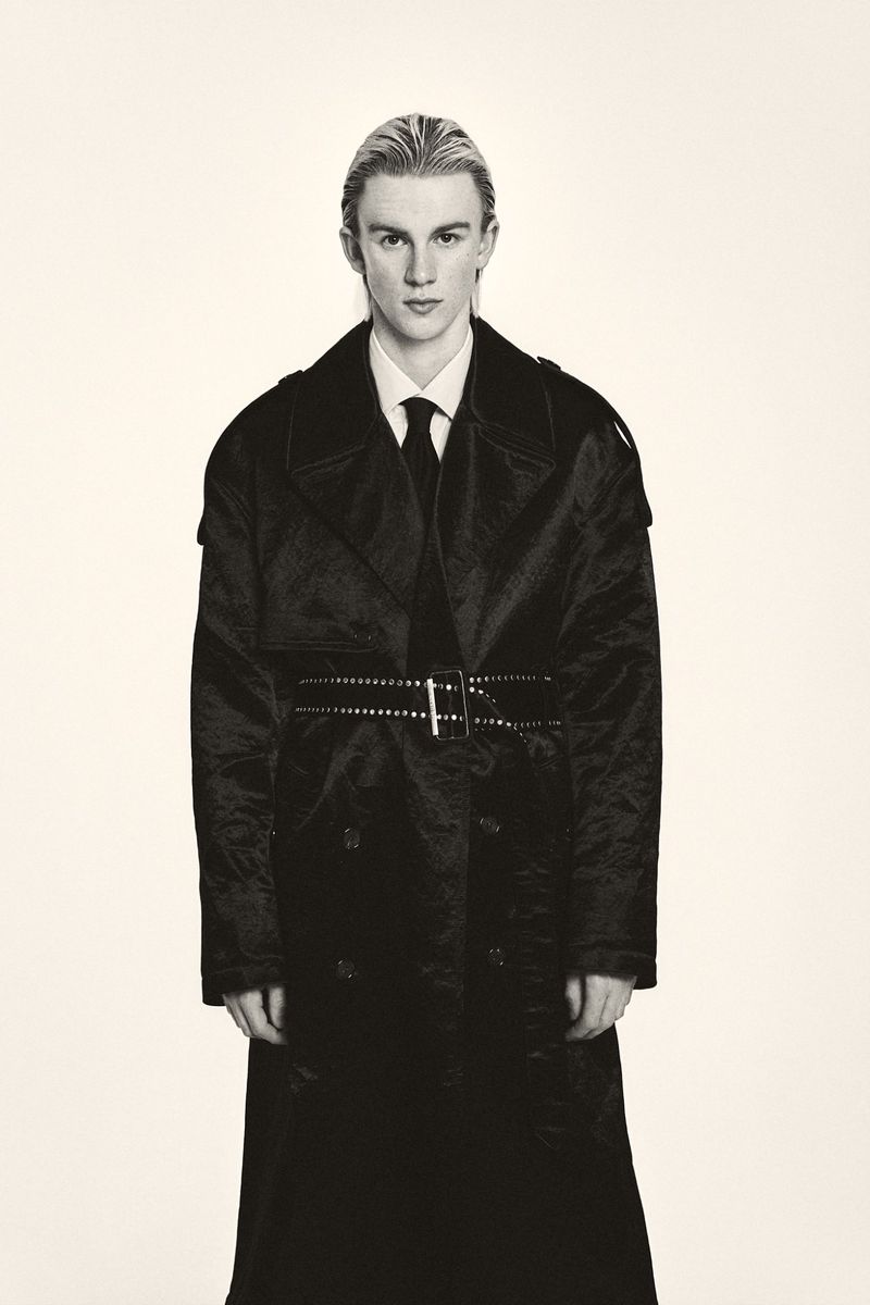 Blonde male model poses indoors wearing a black belted trench coat, white collared shirt, and black tie against an off-white backdrop.