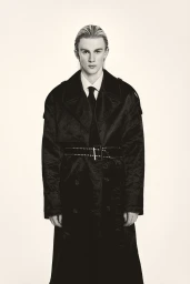 Young male model wearing a dark oversized trench coat and tie against a plain light background