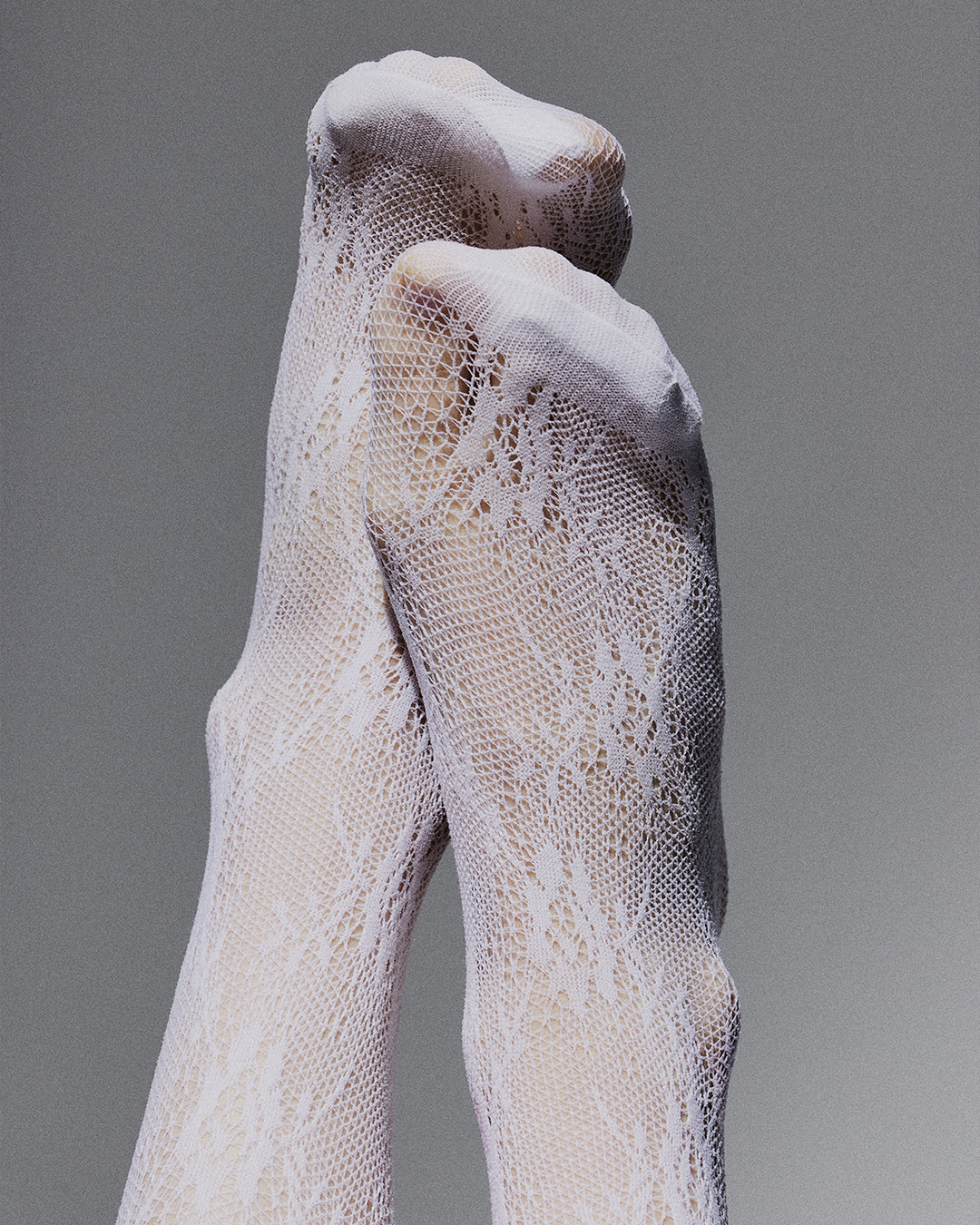 Close-up shot of two legs crossed wearing patterned white stockings, against a grey backdrop in an indoor setting. Fashion.