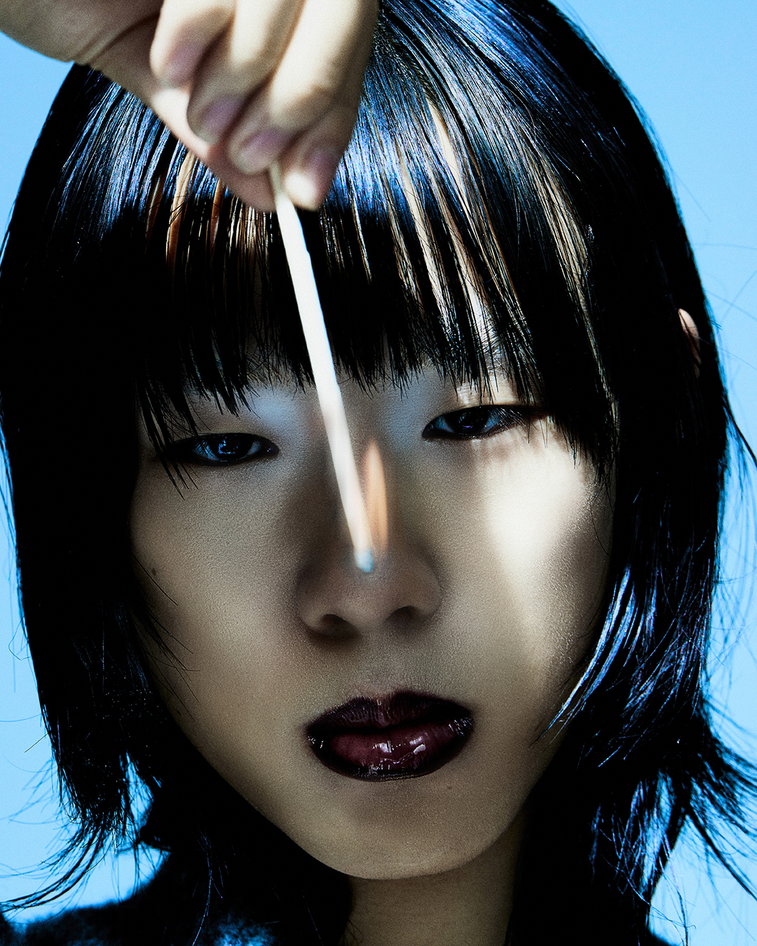 Close-up studio shot of an Asian woman with dark hair, holding a burning match near her face against a pale blue background.