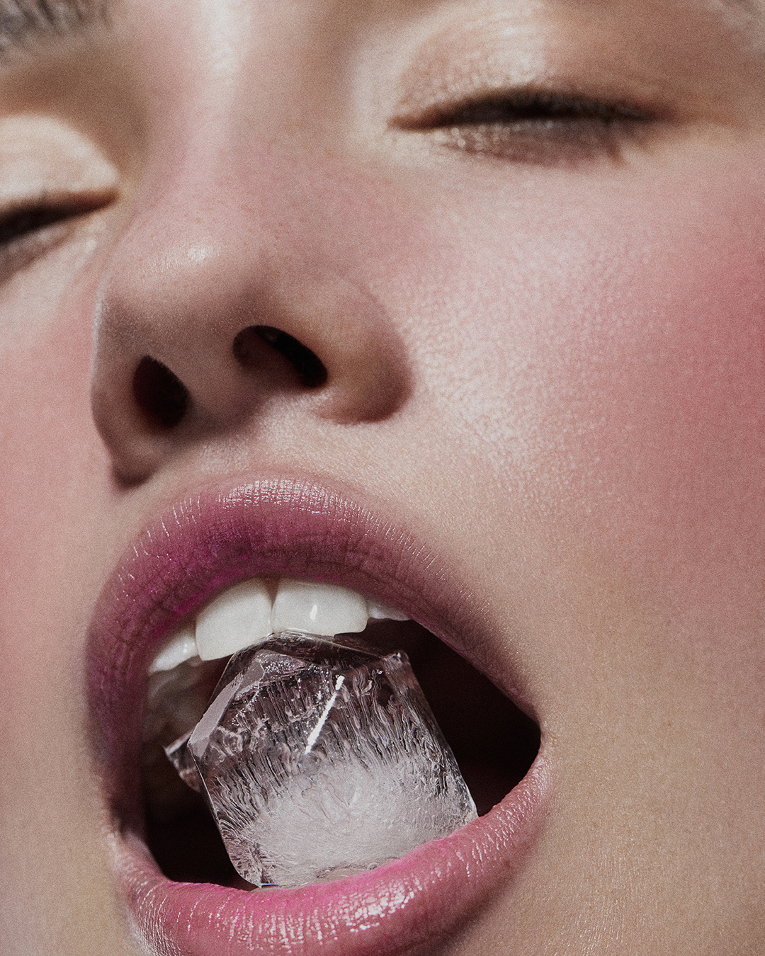 Close-up portrait of a fair-skinned woman with pink lips and eyes closed, holding a clear ice cube in her mouth.