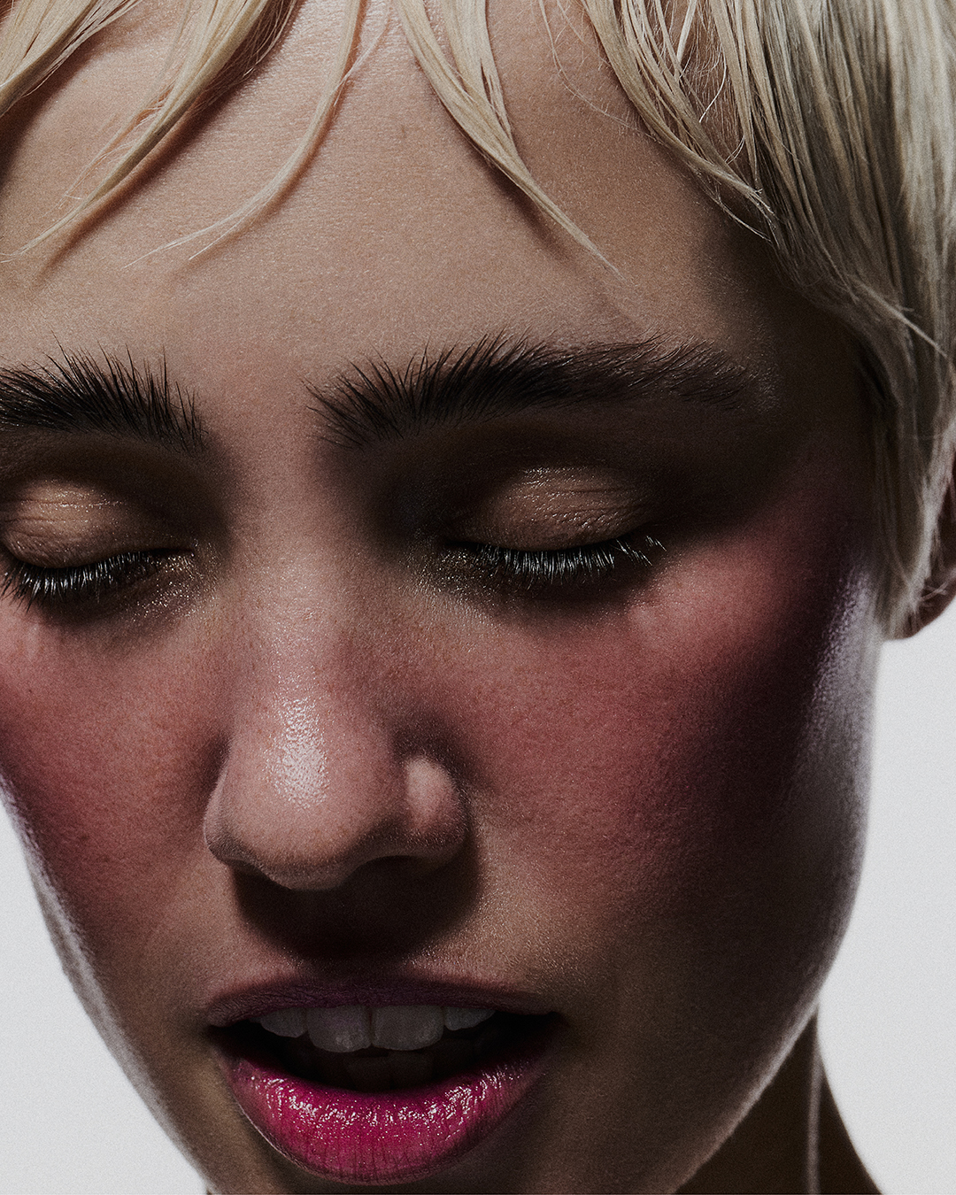 Close-up portrait of a woman with short, blond hair and closed eyes, illuminated by soft lighting that highlights the makeup and skin texture.