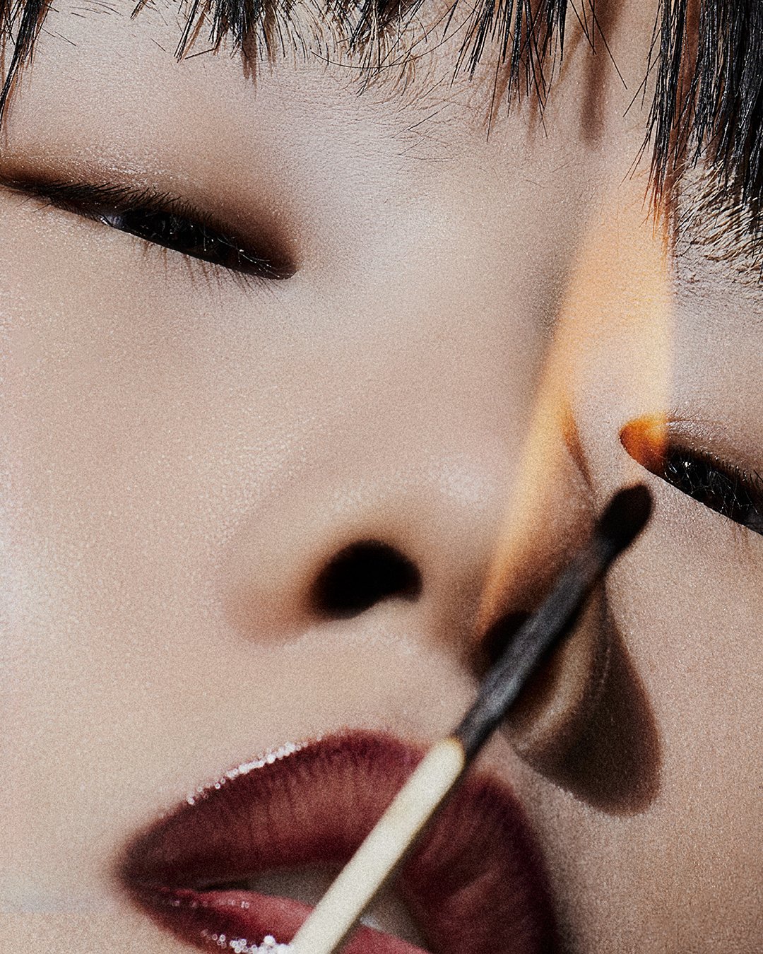 Macro shot of a person's face with dramatic makeup and dark hair, a lit match is positioned near their eye for a daring effect.