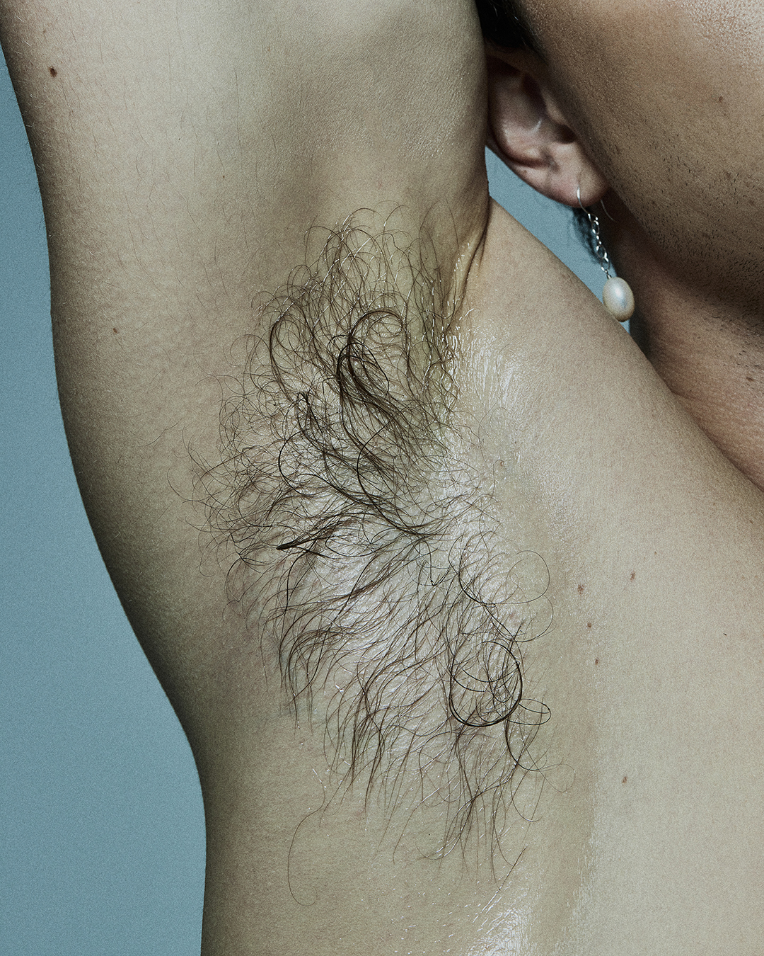 Close-up of a person's armpit with dark hair, skin details and a dangling earring in a well-lit indoor studio setting.