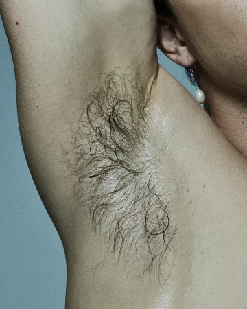 Close-up of a person's armpit with dark hair, skin details and a dangling earring in a well-lit indoor studio setting.