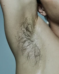 Macro close-up portrait framing a person with a raised arm, showcasing underarm hair and a dangling pearl earring against a pale background