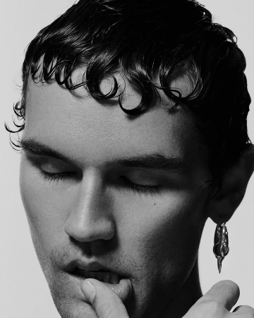 Close-up portrait of a man with wet styled hair and closed eyes, biting his finger against a plain light background