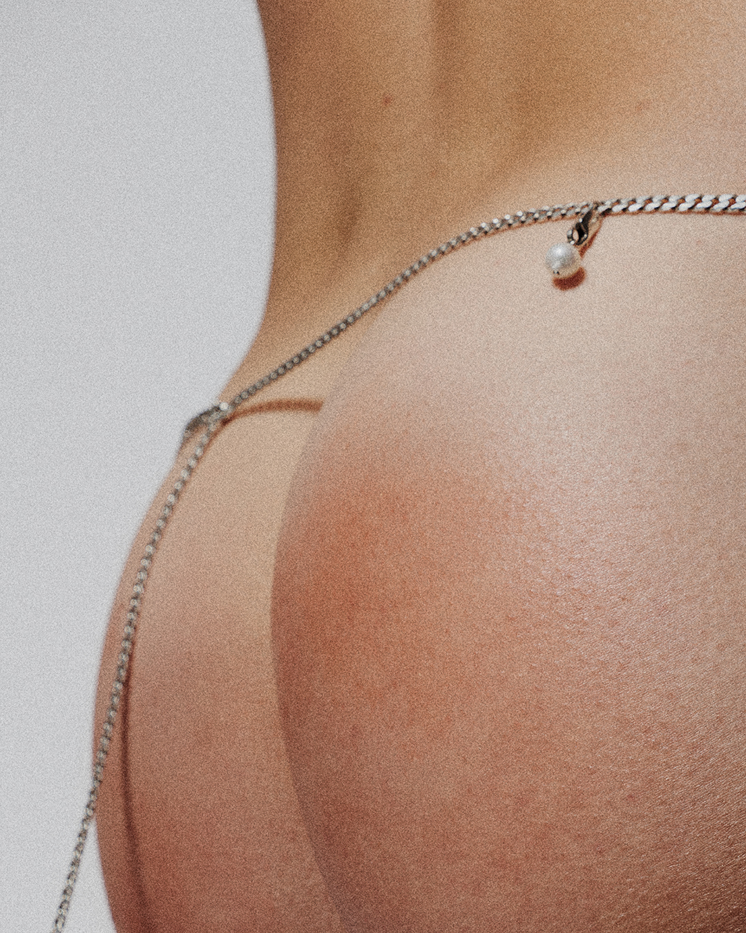 Close-up studio shot of a woman's lower back and buttocks, adorned with a delicate silver chain and pearl detail.