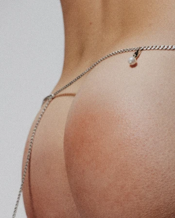 Close-up studio shot of a woman's lower back and buttocks, adorned with a delicate silver chain and pearl detail.