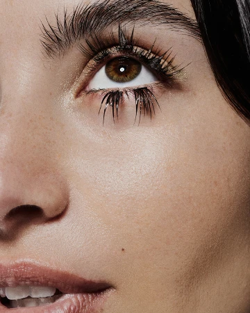 Close-up beauty shot of a woman's face with focus on her eye, showcasing long eyelashes, brown iris, and flawless skin texture in natural light.
