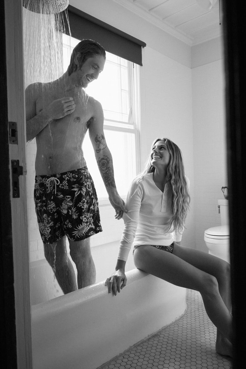 Black and white photo of a smiling couple. A man stands under a shower head while a woman sits on the edge of the bathtub.