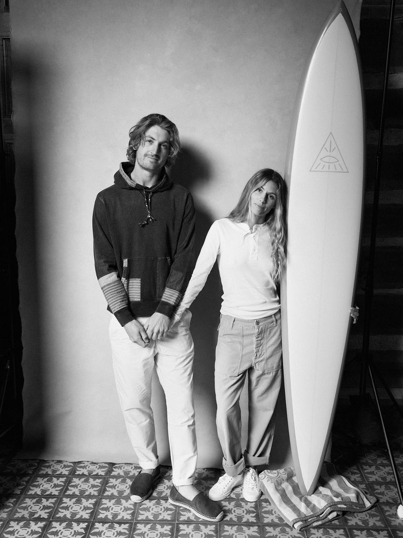 Monochrome shot of a man and woman in casual clothing standing next to a surfboard in a studio setting, captured with natural light.