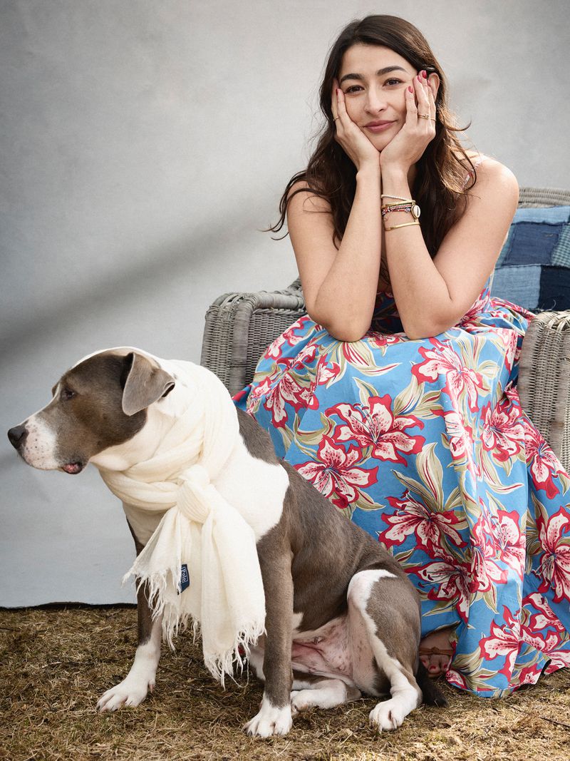 Woman in floral dress poses with a dog wearing a white scarf. The woman is seated with hands on her face and a slight smile.