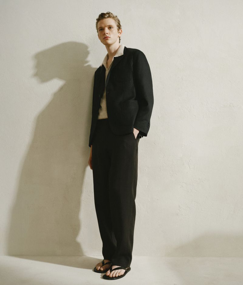 Full-length portrait of an androgynous fashion model posing in black suit against a textured off-white wall in a photo studio.