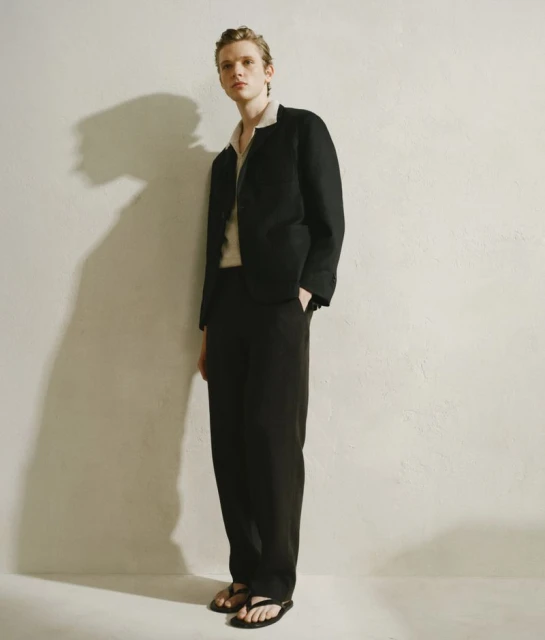 Full-length portrait of an androgynous fashion model posing in black suit against a textured off-white wall in a photo studio.
