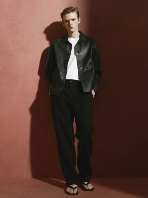 A fair-skinned young adult male poses in black pants, black leather jacket, white T-shirt and black thong sandals against a pink backdrop.