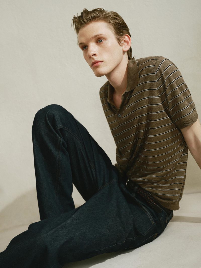 Portrait of a young adult male in a striped polo shirt and jeans, sitting on a neutral surface with soft, diffused lighting.