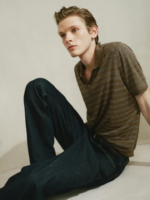 Portrait of a young adult male in a striped polo shirt and jeans, sitting on a neutral surface with soft, diffused lighting.