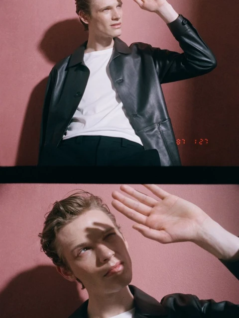 Portrait of a fair-skinned adult male model in a black leather jacket and white t-shirt, shielding eyes, against a pink wall.