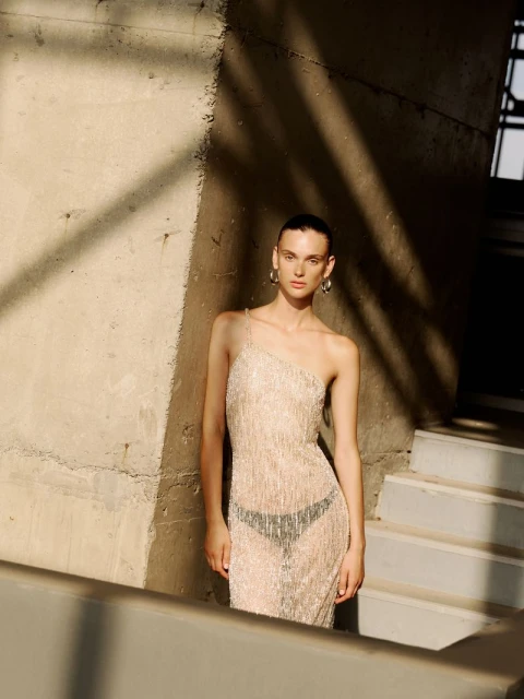 A slender woman posing outdoors near a concrete structure, wearing a shimmering one-shoulder gown with natural light and subtle shadows.