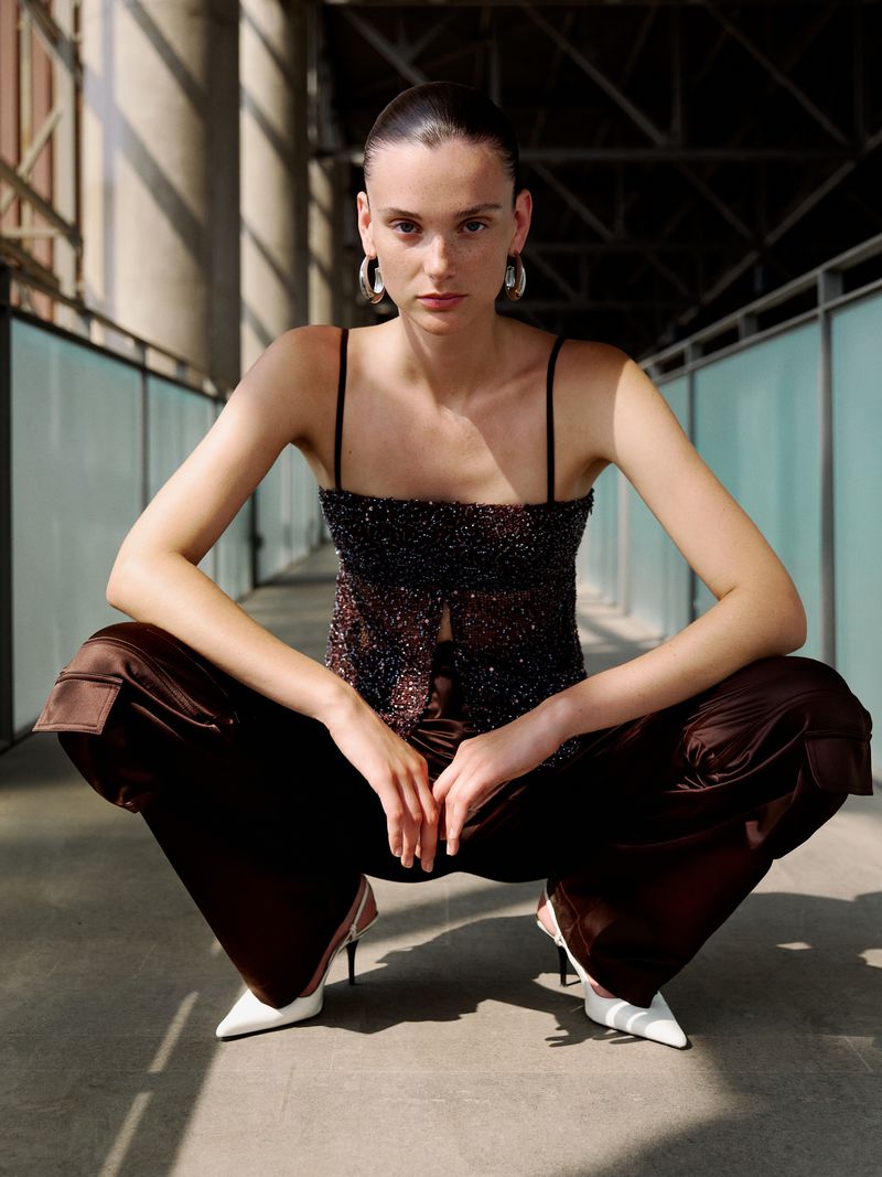 Fashion shot of a slender adult woman with a straight face, in a black beaded top, brown wide-leg pants, and white heels, in a modern space.