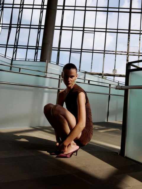 Woman in brown dress and heels strikes a crouching pose indoors with natural light and a large window reflecting the outside sky and building.
