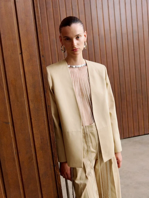 A woman poses in a beige blazer and pantsuit with a fringed top, standing against a wooden wall in a fashion or editorial style photograph.