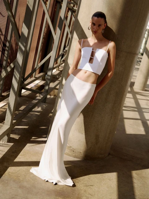 A slender woman poses in a white top and long skirt against an architectural backdrop, casting shadows and strong lines in direct sunlight.