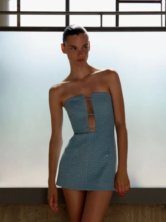 A young woman models a blue strapless mini dress with a cutout detail, posing in a brightly lit indoor setting for a fashion editorial.
