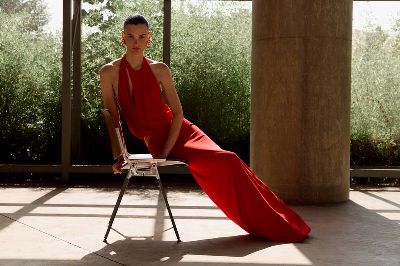 Elegant woman in a vibrant red halter gown sits gracefully on a chair in a sunlit room, set against a lush green backdrop.