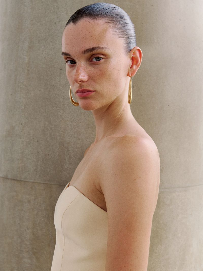 Elegant portrait of a young woman with freckles wearing gold hoop earrings and a strapless top against a neutral concrete wall.