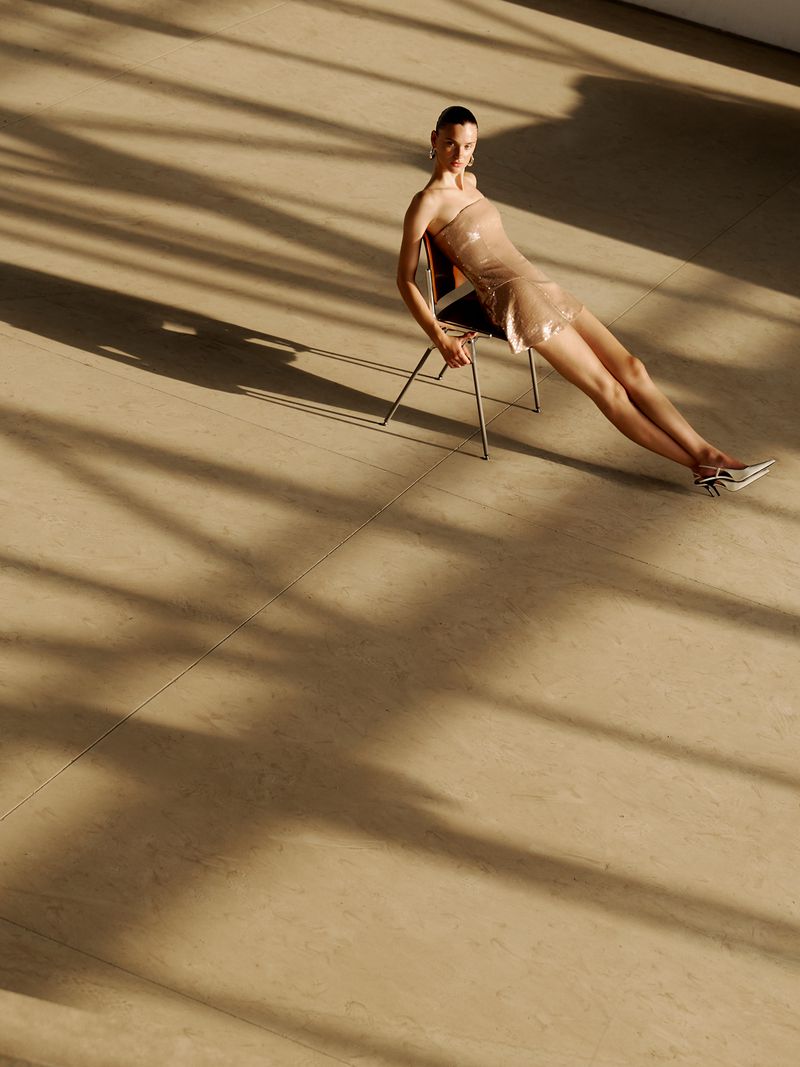 Woman in strapless dress sits on metal chair in light-filled room, casting long shadows on floor. Fashion shot with neutral color palette.
