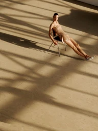 Model seated backwards on a metal chair wearing a strapless metallic dress with dramatic diagonal shadows cast across a concrete floor