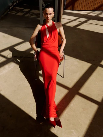 Model wearing a vibrant red halterneck maxi dress seated on a chair under harsh diagonal sunlight shadows.